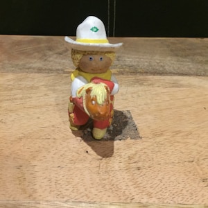 Può includere: Una piccola figurina dipinta a mano di un cowboy a cavallo. Il cowboy indossa un cappello bianco con una fascia gialla e una stella verde, una camicia gialla e pantaloni rossi. Il cavallo è marrone con una criniera e una coda gialle.