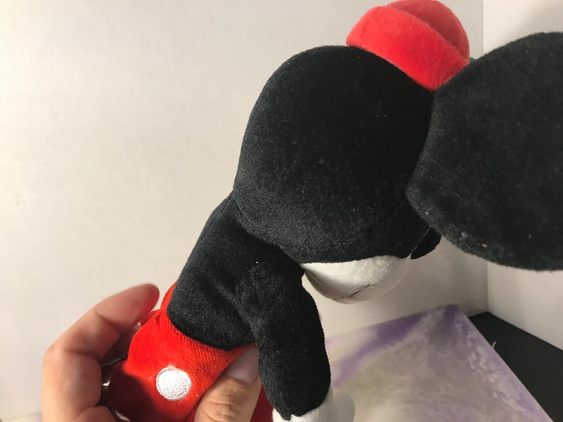 May include: Black and red plush Mickey Mouse toy with a red hat. The toy is in a sitting position with its head tilted down.