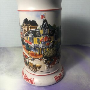 K&ouml;nnte beinhalten: Wei&szlig;e Keramiktasse mit einer bunten Illustration der Main Street, U.S.A. in Walt Disney World. Die Tasse hat einen roten Rand und die Worte "Walt Disney World" und "Disney World" auf dem Boden.