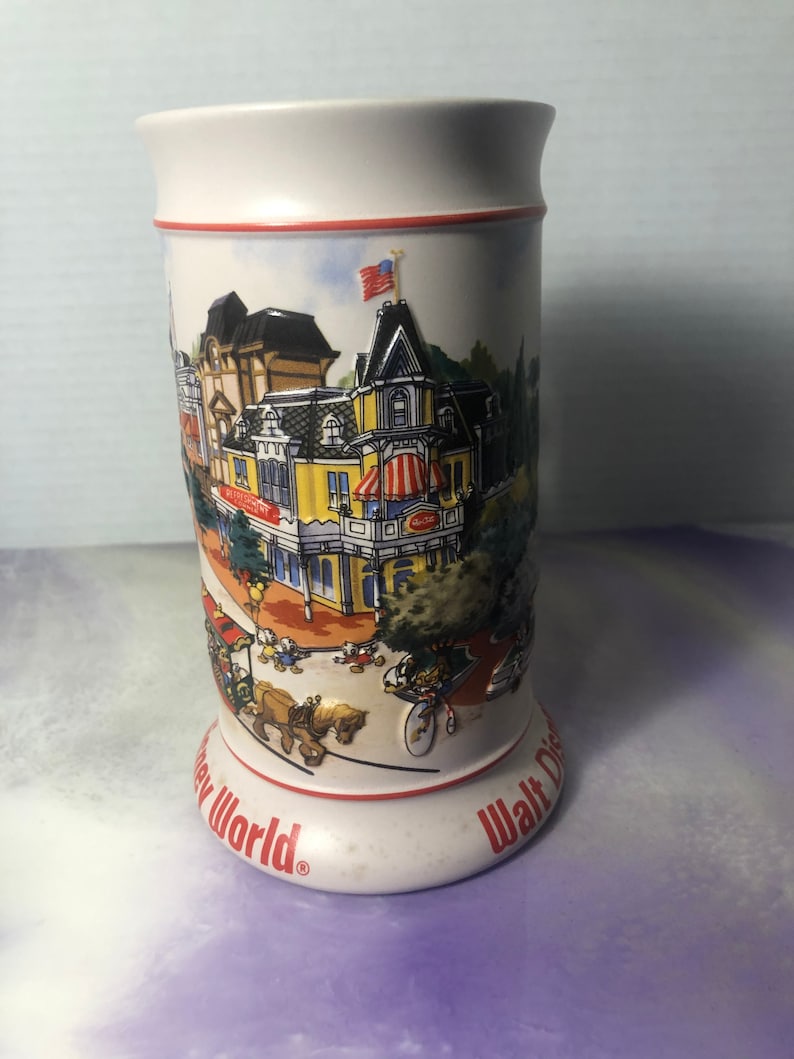 K&ouml;nnte beinhalten: Wei&szlig;e Keramiktasse mit einer bunten Illustration der Main Street, U.S.A. in Walt Disney World. Die Tasse hat einen roten Rand und die Worte "Walt Disney World" und "Disney World" auf dem Boden.