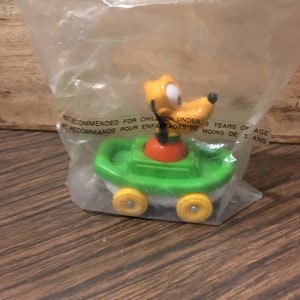 May include: A yellow and orange cartoon dog character in a green toy car with yellow wheels. The toy is packaged in a clear plastic bag. The packaging has a warning label that reads "Not recommended for children under 3 years of age."