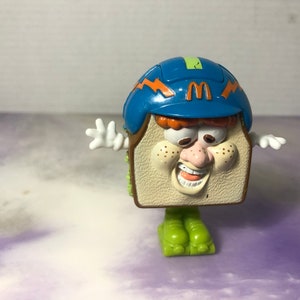May include: A vintage McDonald's toy featuring a sandwich character with a smiling face, freckles, and a blue helmet with the McDonald's logo. The toy has white hands and green feet, and is a collectible item.