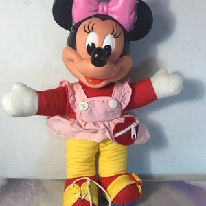 Op de afbeelding: Een pluche Minnie Mouse speelgoed dat een roze jurk met witte stippen, een gele broek en rode schoenen draagt. Ze heeft een roze strik in haar haar en een hartvormig ritsvakje op haar jurk.