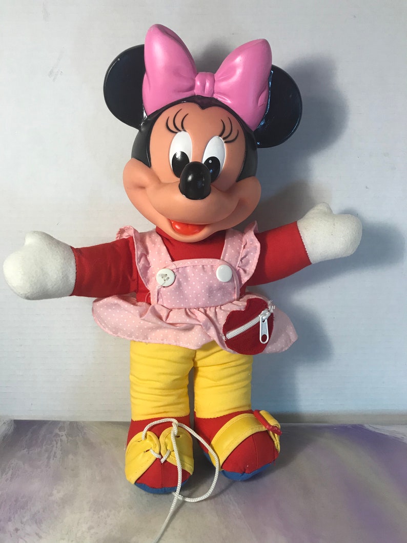 Op de afbeelding: Een pluche Minnie Mouse speelgoed dat een roze jurk met witte stippen, een gele broek en rode schoenen draagt. Ze heeft een roze strik in haar haar en een hartvormig ritsvakje op haar jurk.
