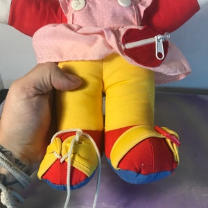 Op de afbeelding: Een pluche pop met een rood shirt, een roze gebloemde jurk, een gele broek en rode en blauwe schoenen. De pop heeft een hartvormig zakje met een rits op de jurk.