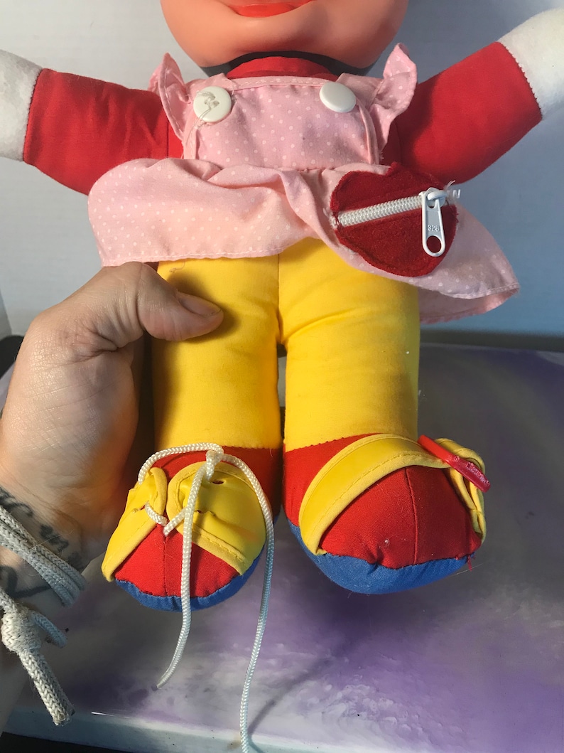 Op de afbeelding: Een pluche pop met een rood shirt, een roze gebloemde jurk, een gele broek en rode en blauwe schoenen. De pop heeft een hartvormig zakje met een rits op de jurk.