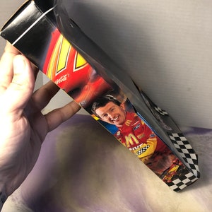 May include: A black and white checkered box with a red, yellow, and black race car on the front. The box features a picture of a race car driver wearing a yellow and black racing suit with the McDonald's logo. The box also has the words "Coca-Cola" and "M&M's" on it.