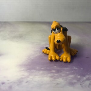 Pode incluir: Uma pequena figura de plástico amarela e preta de um cão de desenho animado com grandes olhos tristes. O cão está sentado em um fundo roxo e branco.