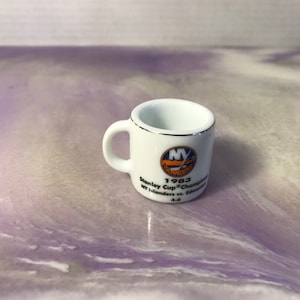 Może przedstawiać: Biały kubek ceramiczny z srebrną obwódką i niebiesko-pomarańczowym logo New York Islanders. Kubek ma napis "1983 Stanley Cup Champions NY Islanders vs. Edmonton 4-0".