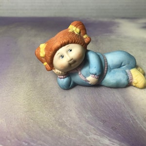 May include: A porcelain figurine of a girl with red hair and yellow bows, wearing a blue and purple jumpsuit. She is lying on her side with her head resting on her hand.