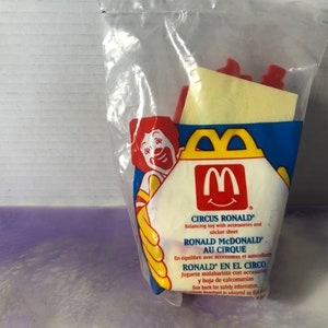 May include: A plastic bag containing a McDonald's Happy Meal toy, Circus Ronald, a balancing toy with accessories and a sticker sheet. The toy features Ronald McDonald in a circus clown costume. The bag is red, yellow, and blue with the McDonald's logo and the text "Circus Ronald", "Ronald McDonald Au Cirque", and "Ronald En El Circo".