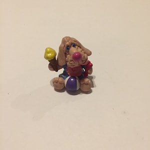 May include: A small plastic figurine of a dog wearing a blue and pink outfit, holding a yellow ice cream cone and a red ball.