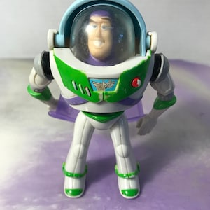 May include: A plastic toy action figure of Buzz Lightyear from the Toy Story franchise. The figure is wearing a white and green spacesuit with a purple and green cape. The figure has a clear plastic helmet with a purple face.