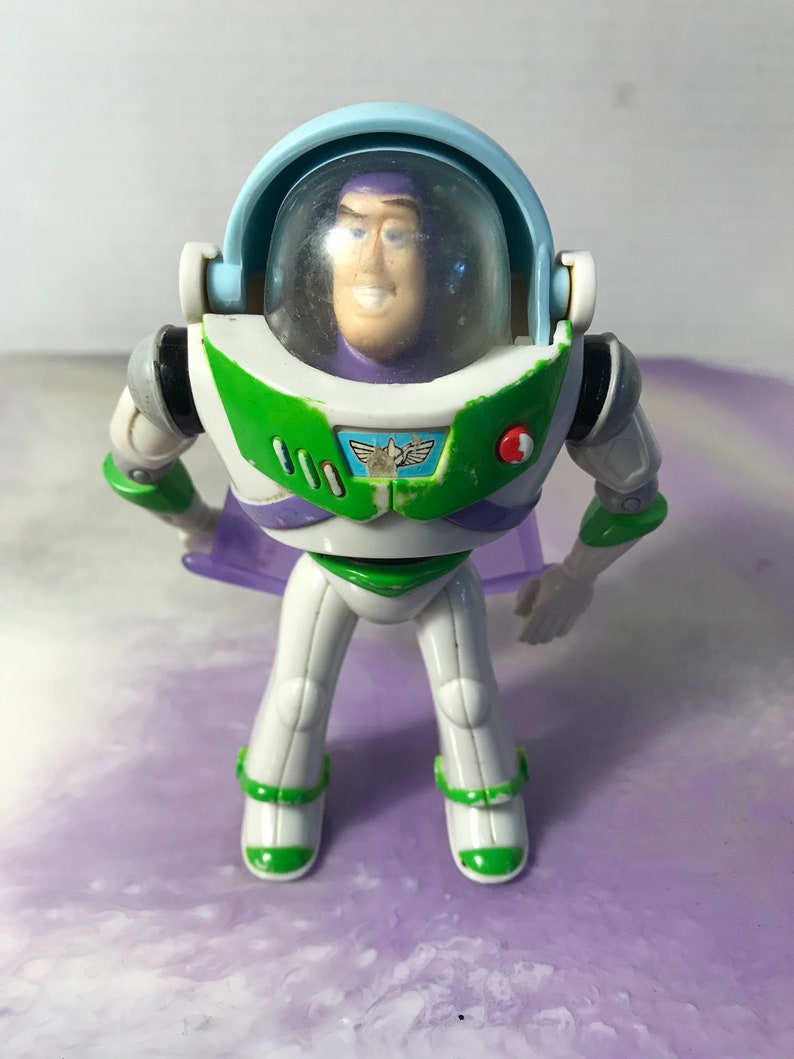 May include: A plastic toy action figure of Buzz Lightyear from the Toy Story franchise. The figure is wearing a white and green spacesuit with a purple and green cape. The figure has a clear plastic helmet with a purple face.