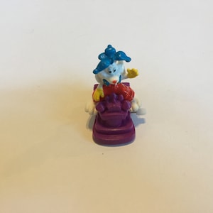 May include: A white rabbit figurine with blue hair and red clothing riding a purple skateboard. The rabbit is holding a yellow object in its right hand.