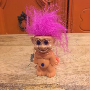 May include: A small plastic troll doll with pink hair and blue eyes. The troll is wearing a blue belly button.