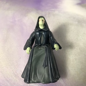 May include: A green-skinned figure wearing a black hooded robe with a grey undergarment. The figure is sitting with its hands resting on its lap.