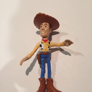 May include: A plastic toy figure of a cowboy, wearing a brown hat, a yellow shirt with a black and white vest, blue pants, and brown boots. The figure has a holster and a red neckerchief. The toy is a character from the animated movie "Toy Story".