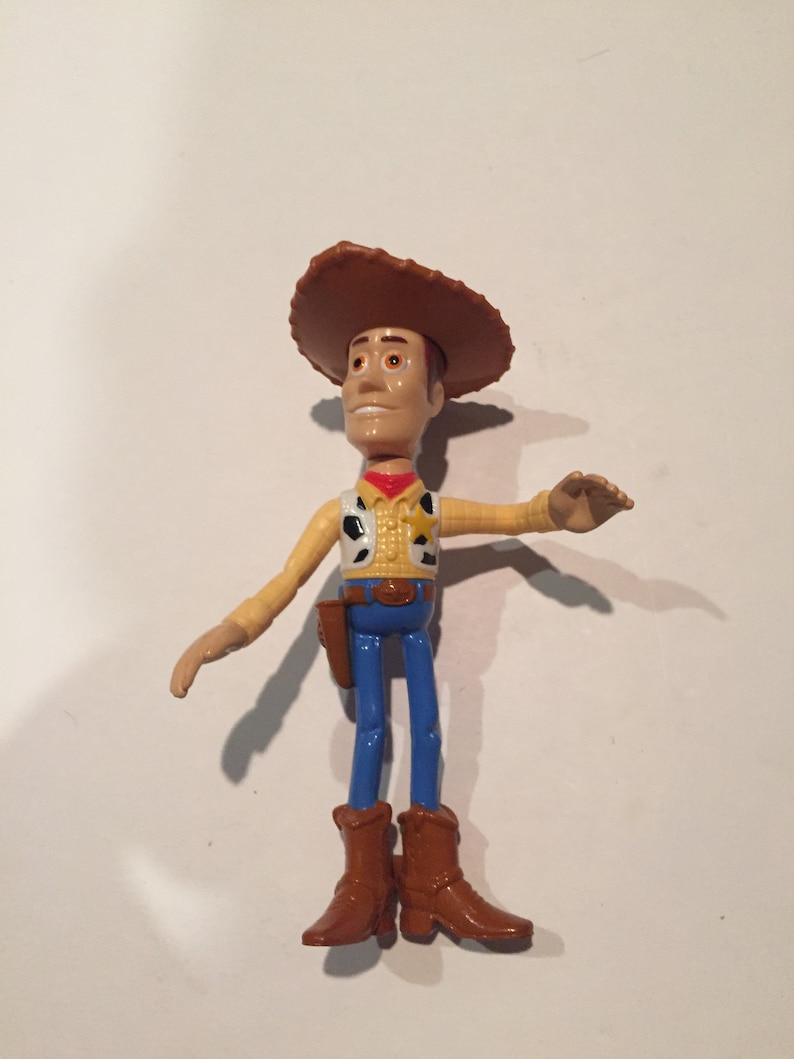 May include: A plastic toy figure of a cowboy, wearing a brown hat, a yellow shirt with a black and white vest, blue pants, and brown boots. The figure has a holster and a red neckerchief. The toy is a character from the animated movie "Toy Story".