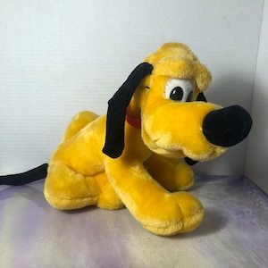 以下が含まれることがあります： 黒い耳と黒い鼻を持つ黄色のぬいぐるみ犬。犬は白い表面に座っています。