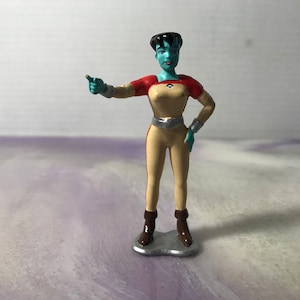 May include: A blue-skinned action figure of a woman wearing a red and tan jumpsuit with a silver belt. She is pointing with her right hand and has black hair.