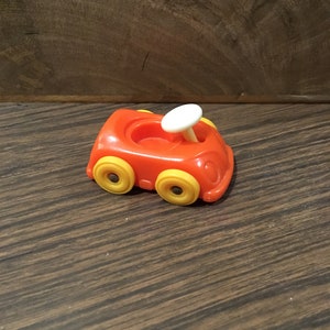 May include: A small, red toy car with yellow wheels and a white steering wheel. The car is made of plastic and has a simple design.