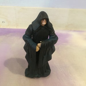 May include: A black hooded figure with a long robe, standing on a purple and white background. The figure has a pale face and is holding its hands together in front of its chest.