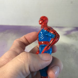 May include: A red and blue plastic action figure of Spider-Man. The figure is posed with its right arm raised and its left arm bent at the elbow.