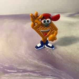 Pode incluir: Uma figura de desenho animado amarela e laranja de um waffle usando um boné de beisebol vermelho e tênis azuis. A figura está sorrindo e acenando com uma mão.