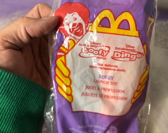 Può includere: Un sacchetto di plastica viola con un logo giallo McDonald's e un'immagine di Goofy. Il sacchetto dice "Disney's Goofy Movie" e "Disney's Extremely Ding" in testo bianco. Il sacchetto dice anche "Bobby Launch Toy" e "Jouet a Propulsion" in testo bianco.