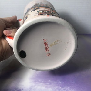 K&ouml;nnte beinhalten: Wei&szlig;e Keramiktasse mit einer bunten Illustration einer Disney-Parade. Am Boden der Tasse steht "DISNEY" in Rot.