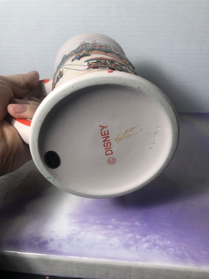 K&ouml;nnte beinhalten: Wei&szlig;e Keramiktasse mit einer bunten Illustration einer Disney-Parade. Am Boden der Tasse steht "DISNEY" in Rot.