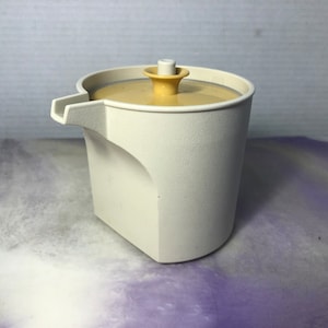 Può includere: Un brocca per panna in plastica bianca con un coperchio beige e un piccolo pomo beige in cima. La brocca ha un beccuccio laterale.