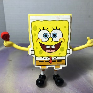 Könnte beinhalten: Gelbe SpongeBob Schwammkopf-Figur mit roter Party-Gabe in der Hand. Die Figur hat schwarze Schuhe, ein weißes Hemd und eine rote Krawatte. Die Figur lächelt und hat die Arme ausgestreckt.