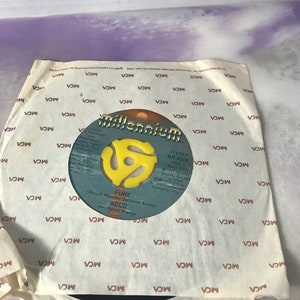 Peut inclure: Un disque vinyle noir avec une étiquette jaune. L'étiquette porte le texte "Millennium" et un logo "A" stylisé. Le disque est intitulé "Funk" par MECO. Le disque est dans une pochette en papier blanc avec un motif répété de "MCA" imprimé dessus.
