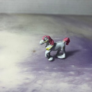 Peut inclure: Un petit cheval en jouet blanc avec des cheveux roses et une garniture dorée autour du cou. Le cheval est debout sur une surface violette et blanche.