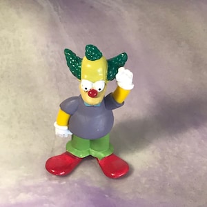 May include: A yellow cartoon figure with green hair, a purple shirt, green pants, and red shoes. The figure is a clown with a red nose and white gloves.
