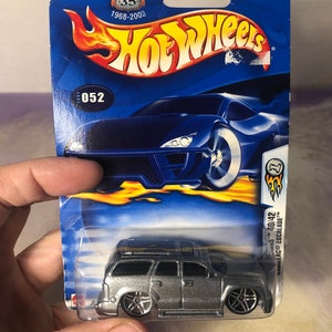 May include: A silver Hot Wheels die-cast toy car of a Cadillac Escalade SUV. The car is on a blue and yellow card with the Hot Wheels logo and the words "Metal Collection" and "56371 3+". The car is number 052 in the series.