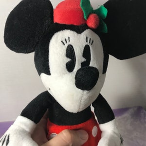 May include: A black and white plush toy of Minnie Mouse wearing a red hat with a green holly sprig and red berry. The toy has a white face with black eyes and a black nose.