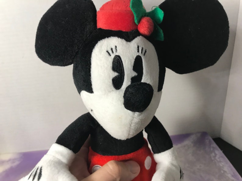 May include: A black and white plush toy of Minnie Mouse wearing a red hat with a green holly sprig and red berry. The toy has a white face with black eyes and a black nose.