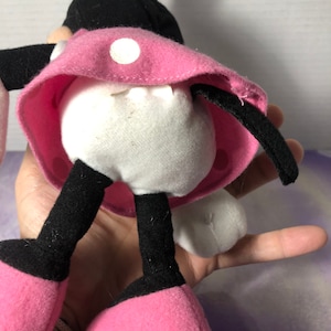 K&ouml;nnte beinhalten: Ein Pl&uuml;sch-Minnie-Maus-Spielzeug mit schwarzen Ohren, einem rosa Kleid mit wei&szlig;en Punkten und schwarzen Schuhen. Das Spielzeug steht auf einer Hand.