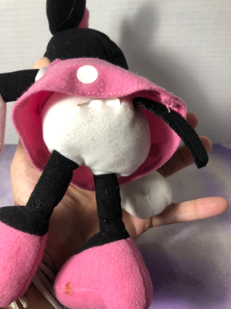 K&ouml;nnte beinhalten: Ein Pl&uuml;sch-Minnie-Maus-Spielzeug mit schwarzen Ohren, einem rosa Kleid mit wei&szlig;en Punkten und schwarzen Schuhen. Das Spielzeug steht auf einer Hand.