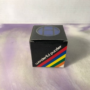 Może przedstawiać: Czarne pudełko z niebieską kostką do układania w środku. Na pudełku widnieje napis "wonderful puzzler" w kolorze czerwonym, białym, niebieskim, żółtym i zielonym.