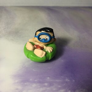 May include: A small plastic figurine of a boy wearing a blue diving mask and red swim trunks, sitting in a green inflatable ring.