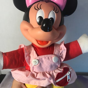 Op de afbeelding: Een pluche Minnie Mouse pop die een roze en wit geblokt jurkje draagt met een rood hartvormig zakje. Ze heeft een roze strik in haar haar en een brede glimlach.
