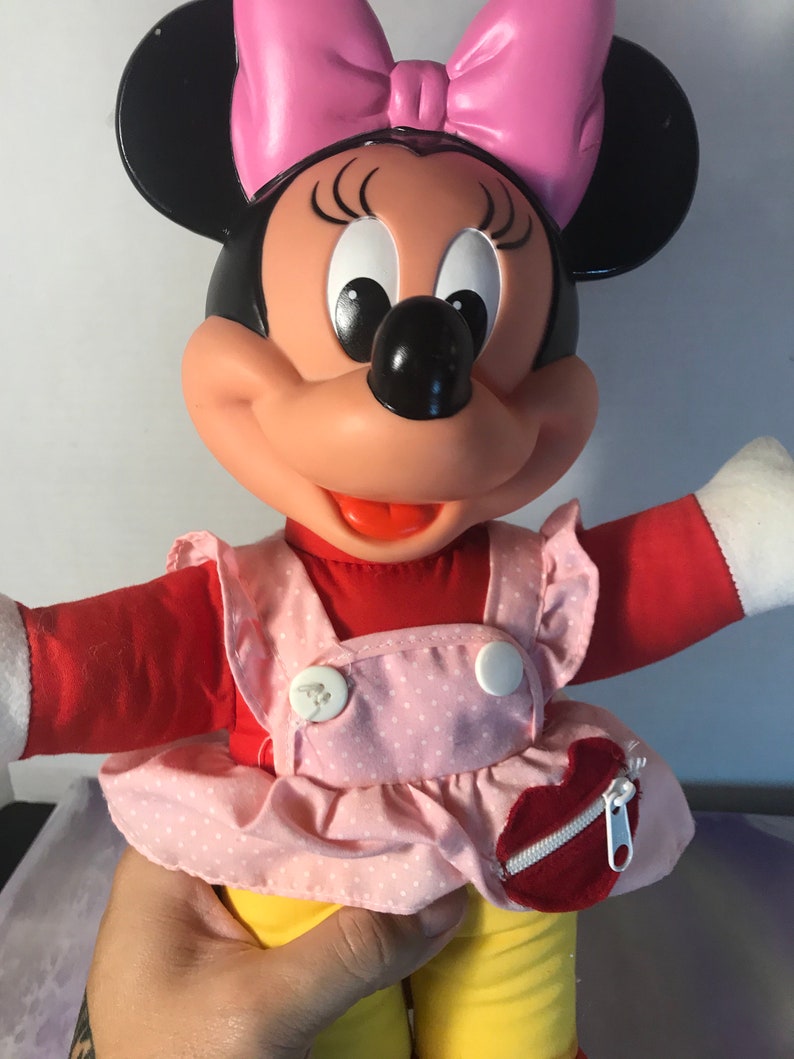 Op de afbeelding: Een pluche Minnie Mouse pop die een roze en wit geblokt jurkje draagt met een rood hartvormig zakje. Ze heeft een roze strik in haar haar en een brede glimlach.