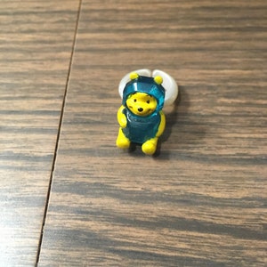 以下が含まれることがあります： 青いフード付きの衣装を着た、小さな黄色のくまのプーさんのフィギュア。フィギュアはプラスチック製で、漫画のような外観をしています。