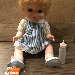 May include: A vintage pull-string doll with blonde hair, blue eyes, and a blue and white striped dress. The doll is sitting on a wooden surface and holding a clear plastic baby bottle with a pink nipple. The doll has a tag that reads "Pullan Doll Canada's Finest".