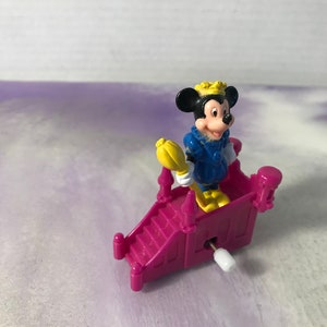 May include: A wind-up toy featuring Mickey Mouse dressed as a king, standing on a pink castle-shaped platform with a yellow scepter.