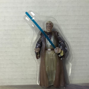 May include: A plastic-packaged action figure of a Star Wars character, Obi-Wan Kenobi, holding a blue lightsaber. He is wearing a brown robe and has a white beard.